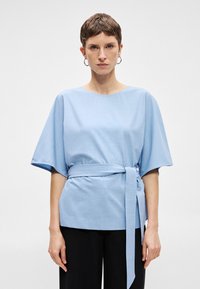 Ανοιχτό μπλε μπλούζα από υφασμένο ύφασμα, με κοντά μανίκια, στρογγυλή λαιμόκοψη και ζώνη στη μέση που δένει για ρυθμιζόμενη εφαρμογή.