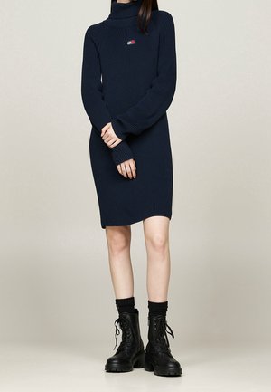 Kobieta ubrana w granatową prążkowaną sukienkę z golfem, zestawioną z czarnymi masywnymi sznurowanymi botkami i czarnymi skarpetkami na jednolitym tle.