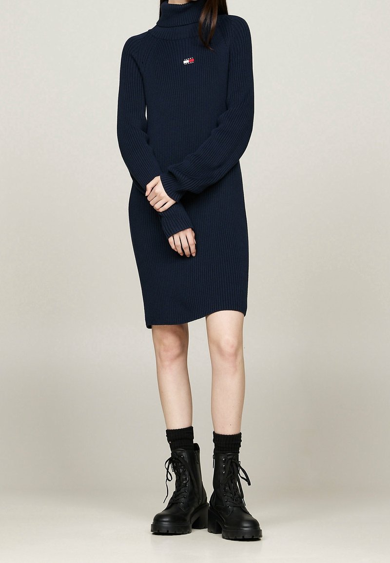 Femme portant une robe-pull à col roulé côtelée bleu marine associée à des bottes noires épaisses à lacets et des chaussettes noires, sur un fond uni.