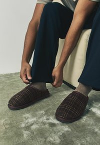 Braune Karoschlafsäcke mit weicher Textur und gepolsteter Sohle, getragen mit grauen Socken und blauen Hosen, auf einem strukturierten Boden.