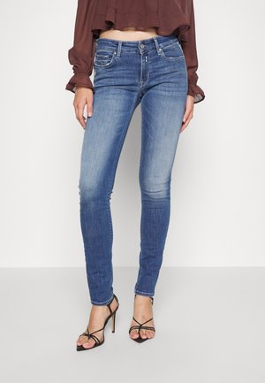 Frau trägt mittelhohe, ausgewaschene blaue Skinny-Jeans, eine bordeauxfarbene, kurze Bluse mit Puffärmeln und schwarze Riemchensandalen mit hohen Absätzen.