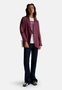 Femme portant un blazer violet, un t-shirt graphique blanc, un jean bleu foncé, des sandales argentées et portant un petit sac à bandoulière blanc.