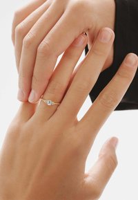 Anillo de oro con una gema azul central, rodeada de pequeñas piedras claras, montado en una delicada banda. Manos entrelazadas.