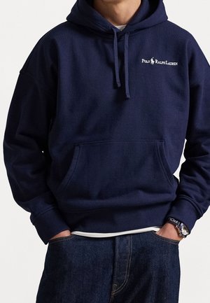 Homme portant un sweat à capuche Polo Ralph Lauren bleu marine avec poche avant et un jean bleu foncé, mains dans les poches, et une montre au poignet gauche.