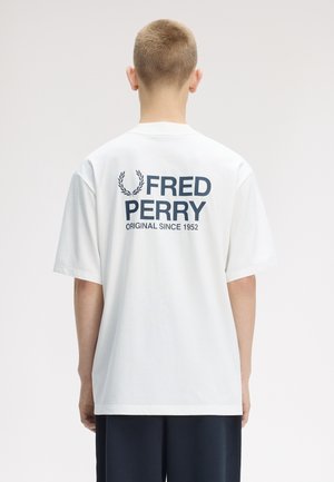 Personne aux cheveux courts portant un t-shirt blanc Fred Perry avec un texte et un logo bleu marine, debout de dos sur un fond uni.