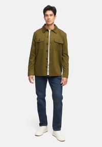 Olivenfarvet skjortejakke med to brystlommer, sorte knapper og krave, parret med blå jeans og hvide sneakers.