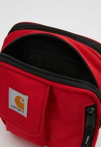 Red fabric bag with black zippers, featuring a front pocket and a Carhartt logo patch. The interior is dark and smooth-textured.