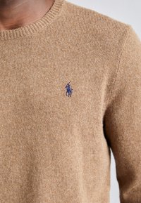 Pull en tricot beige avec un col ras du cou côtelé et un petit logo brodé violet sur la poitrine gauche ; tissu texturé avec une finition douce.