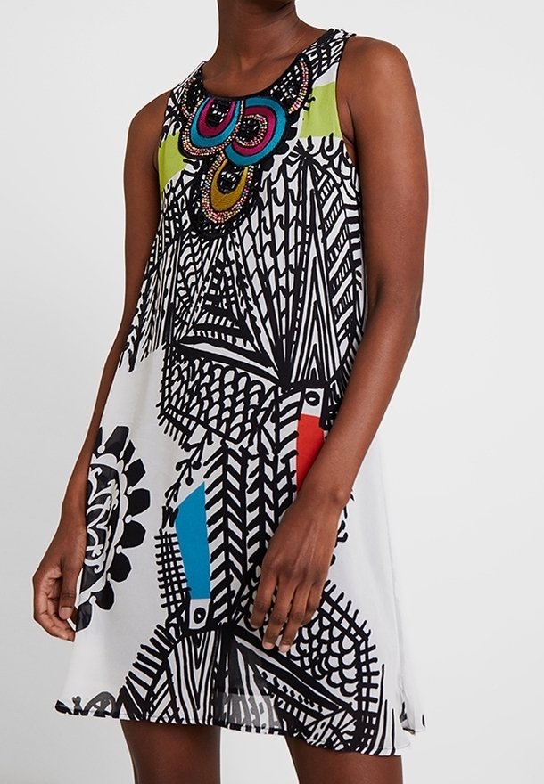 Robe blanche sans manches avec des motifs géométriques et abstraits noirs audacieux, avec un collier coloré bleu, rouge, vert et orné de détails.