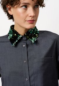 Donkergrijze blouse met een pailetten kraag in groene, zwarte en blauwe tinten. Sluiting met knopen en een borstzak zijn zichtbaar.