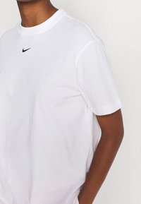T-shirt bianco in cotone con scollatura rotonda, maniche corte e piccolo logo Nike nero centrato vicino al collo. Texture morbida.