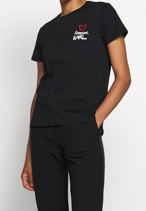 Persoon draagt een zwart T-shirt met een rood hart en witte tekst "FOREVER KARL" op de borst, gecombineerd met een zwarte broek.