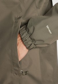 Jachetă verde măslinie cu un buzunar cu fermoar, manșete elastice și logo-ul „DRYVENT”. Materialul pare neted cu un finisaj mat.