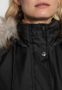 Zwarte parka met een capuchon van nepbont, voorzien van een hoge kraag en drukknoopsluitingen. Zachte textuur en stevig materiaal zichtbaar.