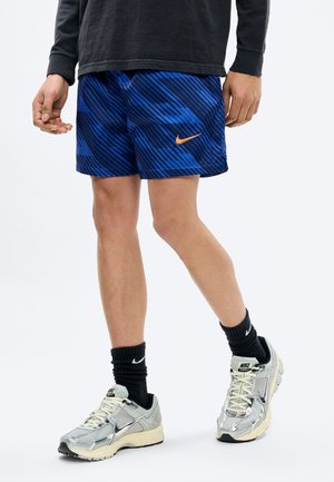 Persoon die blauwe Nike-shorts met patroon, zwarte Nike-sokken en zilveren Nike-sneakers draagt, staand tegen een effen achtergrond.