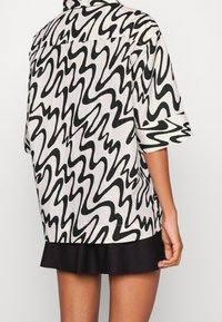 Camicia a maniche corte bianca con un motivo ondulato nero, caratterizzata da un fit rilassato e colletto con bottoni. Una gonna nera spunta da sotto.