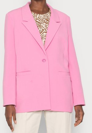 Personne portant un blazer rose vif à un bouton sur un haut imprimé zèbre marron et blanc, et un pantalon blanc, sur un fond uni.