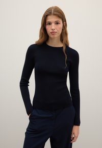 Top noir côtelé à manches longues avec un col rond, présentant une silhouette ajustée et une texture lisse, associé à un pantalon foncé.