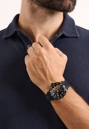 Muškarac u tamnoplavoj teksturiranoj polo majici podešava ovratnik lijevom rukom, na zapešću crno-narančasti analogni ručni sat.