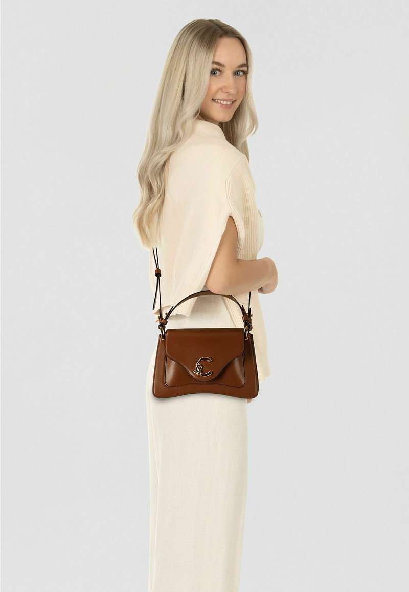 Bruine leren handtas met een gebogen ontwerp, zilverkleurige hardware en een opvallend logo. Het heeft een verstelbare band en een gladde textuur.