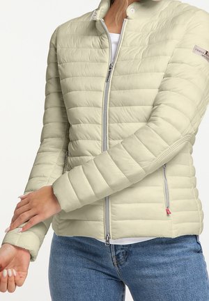 Personne portant une veste matelassée beige clair avec une fermeture éclair à l'avant et un jean bleu, ajustant la manche du bras gauche.