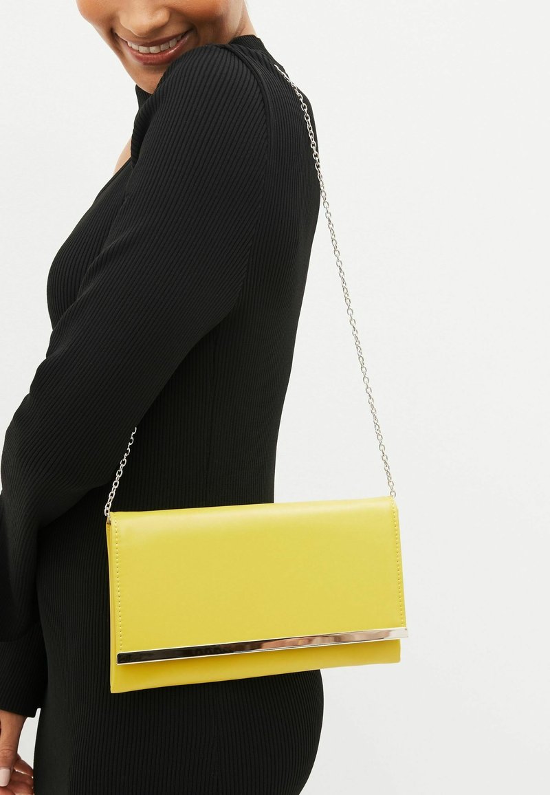 Femme en robe noire portant un sac rectangulaire jaune avec une bandoulière en chaîne argentée et un accent métallique à l'avant.