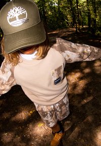 Cappello khaki con logo, felpa beige con maniche camouflage, pantaloni corti con fantasia e stivali gialli. Ambientazione naturale con vegetazione sullo sfondo.