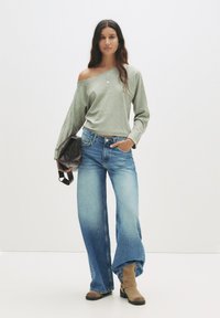 Helles graues Off-Shoulder-Pullover kombiniert mit hochgeschnittenen, weit geschnittenen blauen Jeans und tanfarbenen Wildleder-Ankle Boots, während sie eine braune Tasche hält.