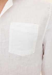 Camicia bianca in lino con tasca sul petto, tessuto strutturato e colletto classico. Il design complessivo è semplice e minimalista.