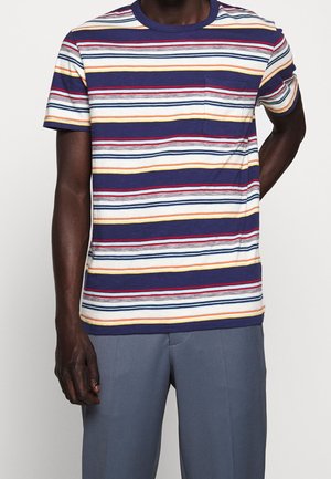 Mężczyzna ubrany w granatowo-białą, czerwoną i żółtą pasiastą koszulkę z krótkim rękawem i kieszonką na piersi oraz szare spodnie, stojący na jednolitym tle.