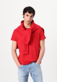 Rode katoenen T-shirt met korte mouwen, gecombineerd met een rode trui om de hals gedrapeerd. Model draagt lichtblauwe denim jeans.