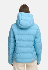 Ljusblå puffjacka med en plyschhuva, quiltat mönster och elastiska ärmslut. Har en diskret logotypdekoration nära fållen.