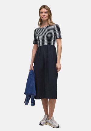 Femme portant une robe noire et blanche à rayures, tenant une veste en denim bleu, debout, portant des baskets argentées et blanches.
