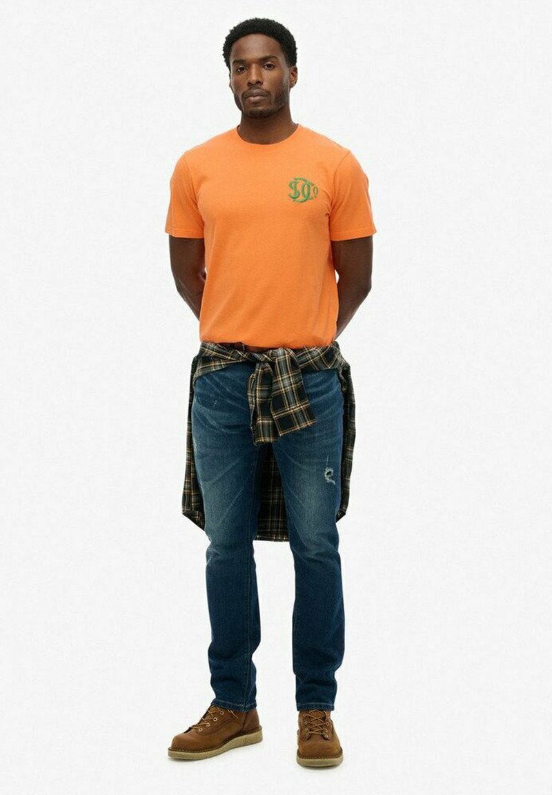 Camiseta naranja con logo verde, jeans azules con detalle desgastado y una camisa de cuadros anudada en la cintura. Botas marrones completan el look.