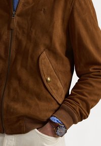 Jachetă bomber din suede maro, cu închidere cu fermoar, manșete redate și un buzunar lateral cu un buton din alamă. Pur tată peste o cămașă albastră.