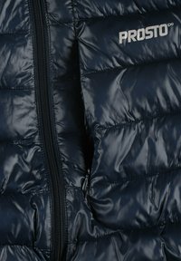 Granatowa kurtka puchowa wykonana z błyszczącego, pikowanego materiału; posiada pełny zamek błyskawiczny i srebrne logo "PROSTO" na przodzie.