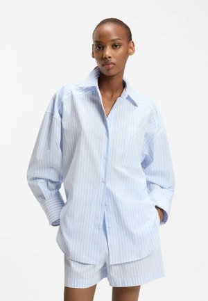 Frau trägt ein hellblau-weiß gestreiftes, übergroßes Hemd mit Knöpfen und passende Shorts, steht mit den Händen in den Taschen.