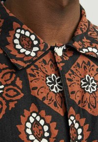 Camisa de manga corta en negro con un patrón geométrico floral en naranja y blanco. Presenta un cuello puntiagudo y un cierre frontal de botones.
