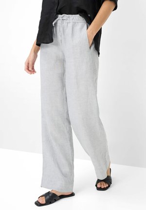 Persoon die lichtgrijze linnen wijde broek met elastische taille en zwarte sandalen draagt, staand met één hand in de zak.