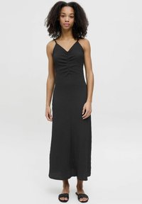 Abito maxi nero con spalline sottili, tessuto texturizzato, corpino arricciato e una silhouette fluida. Abbinato a sandali neri con fascia.
