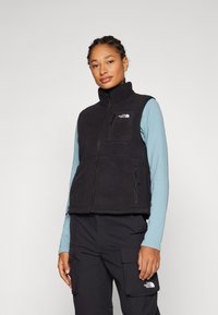 Black fleece vest with high collar, full front zipper, and two side zip pockets. Features a small chest pocket with logo detail.