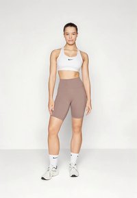 Reggiseno sportivo bianco con logo Nike nero, abbinato a pantaloncini a vita alta color marrone chiaro. Modello con calzini e scarpe bianchi. Sfondo semplice.