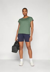Grön kortärmad träningsskjorta kombinerad med marinblå shorts. Modellen håller en svart gymväska; bär vita sneakers med texturerade sulor.