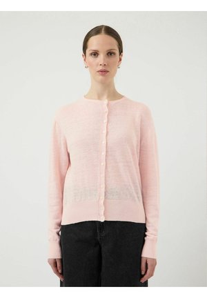 Mujer con un cárdigan abotonado de color rosa claro y pantalones negros, de pie frente a un fondo blanco liso, mirando hacia adelante.