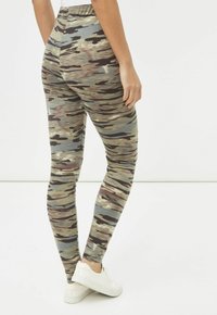 Persona vistiendo leggings ajustados con un patrón de camuflaje y zapatillas blancas, de pie con un pie ligeramente levantado frente a un fondo liso.