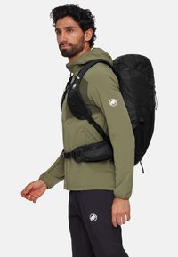Svart ryggsäck med texturerad yta, justerbara remmar och en zippered midjeväska; bärs av en person i en långärmad olivgrön skjorta.