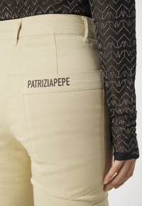 Beige pants with a smooth texture, featuring a back pocket with "PATRIZIAPEPE" printed in dark brown. Sheer black top with a zigzag pattern.