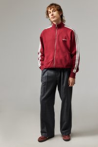 Punane üles tõmmatav jakk valgete triipudega varrukatel, hallid volditud püksid ja maroonid loafersid. Kangas tundub pehme ja tekstuuriga.