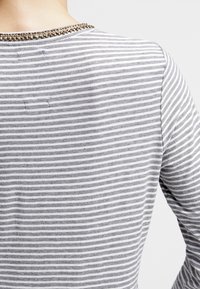 Persona vestida con una camiseta de manga larga a rayas horizontales en gris y blanco, con un cuello de cuentas, vista desde la espalda y el hombro.