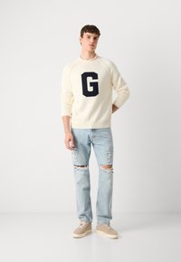 Cream sweater with a navy "G" logo, ribbed cuffs, worn with light wash distressed jeans, and beige sneakers. Simple, casual outfit.